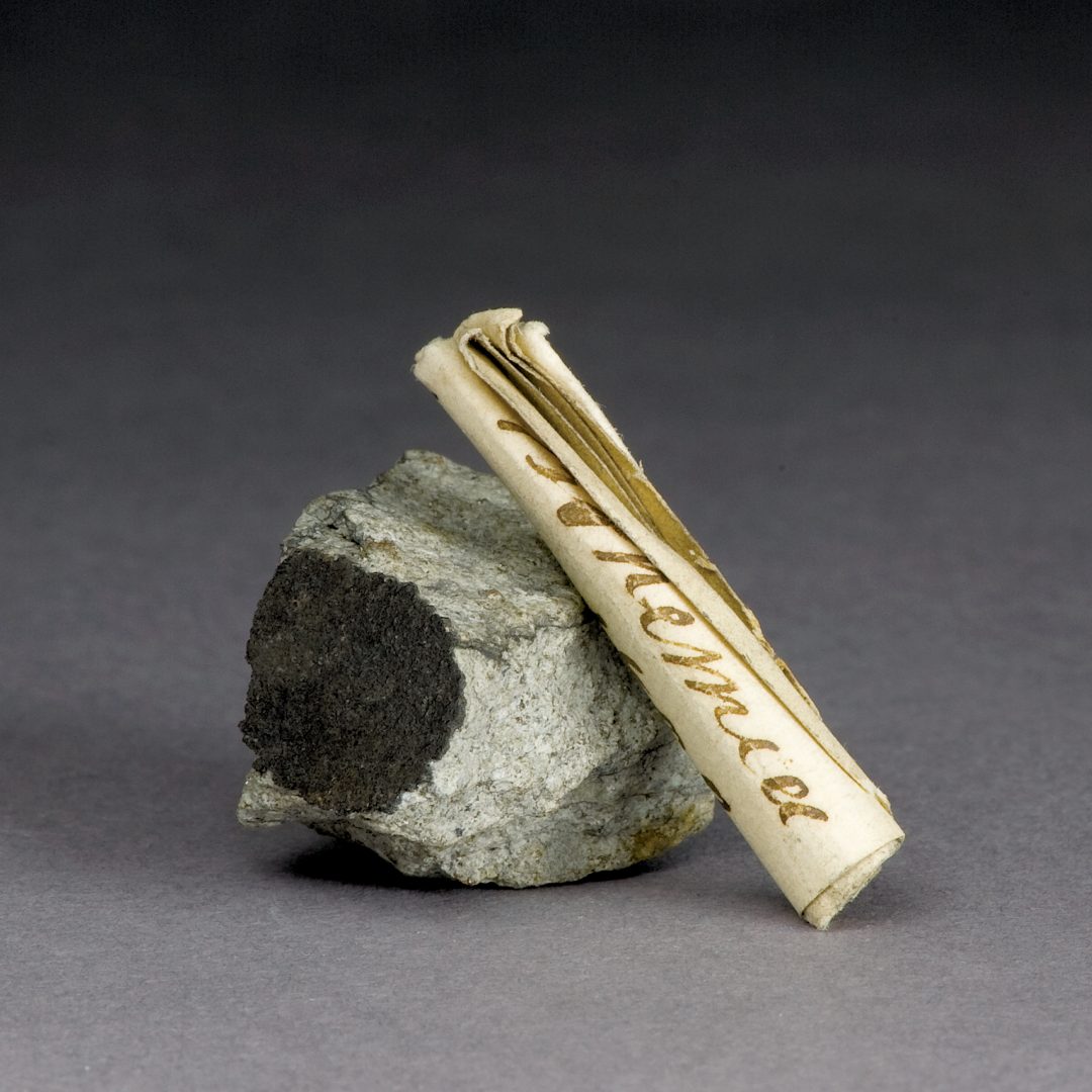 A tightly rolled parchment scroll is propped against a light grey rock with a patch of black fusion crust.